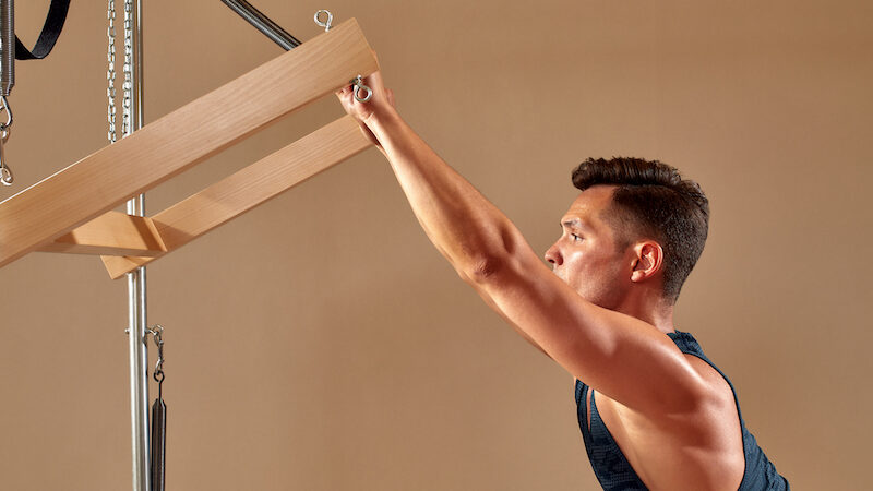 handsome pilates male instructor performing stretching balance fitness exercise on small barrel equipment, at the pilates studio modern onterior indoor.